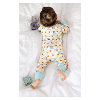 Baby lying on a white blanket wearing a colorful onesie with toys around.