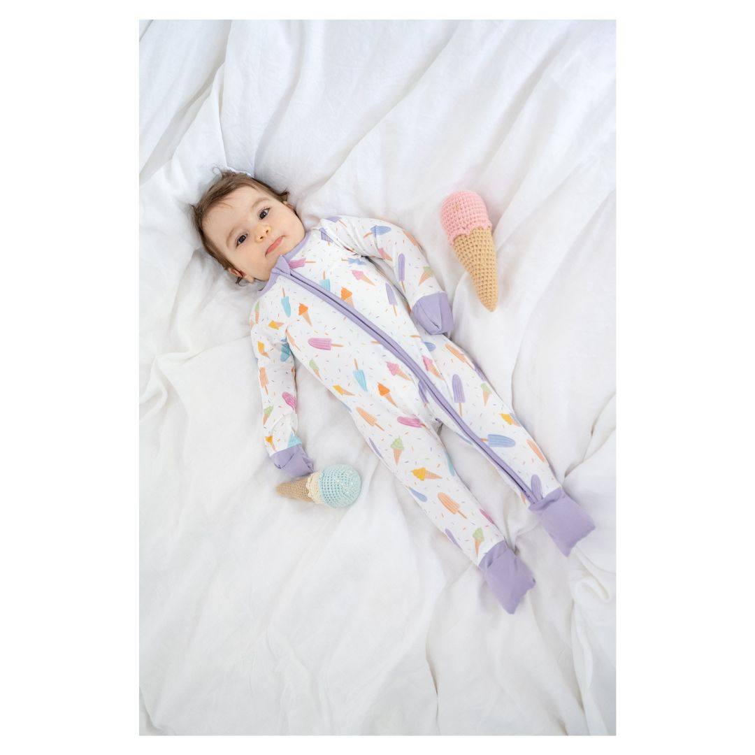 Baby in a colorful onesie lying on a white blanket with ice cream toy.