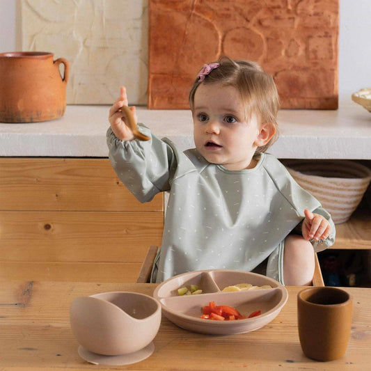 An olive green long-sleeved waterproof bib apron for babies by Trois Kilos Sept, featuring a front pocket for catching food.